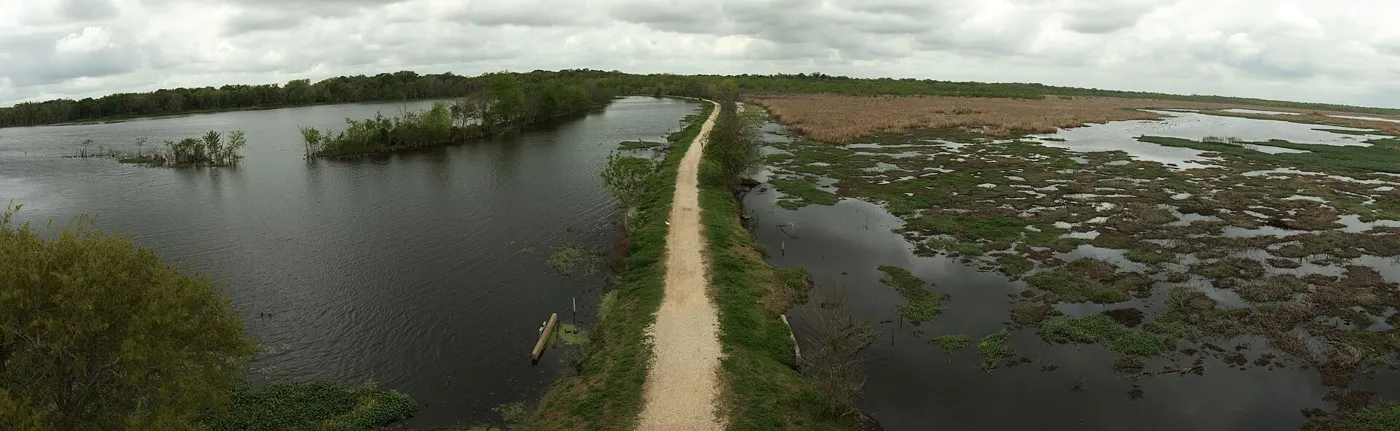 A dirt levee trail running between two marshy lakes under a cloudy sky, seen from above