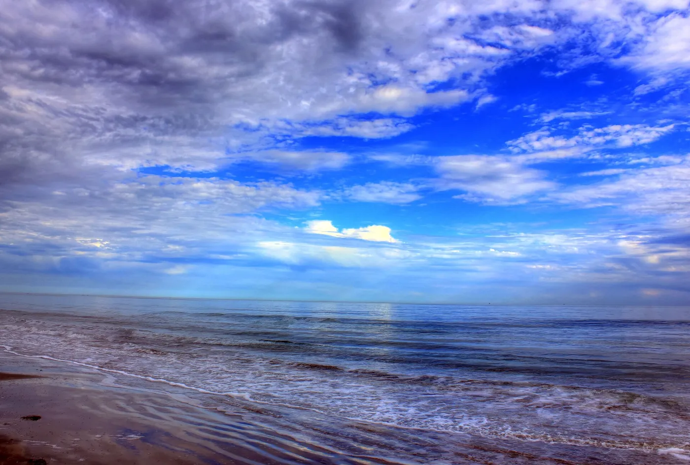 Waves rolling onto a flat Gulf beach under a sky full of white and blue clouds