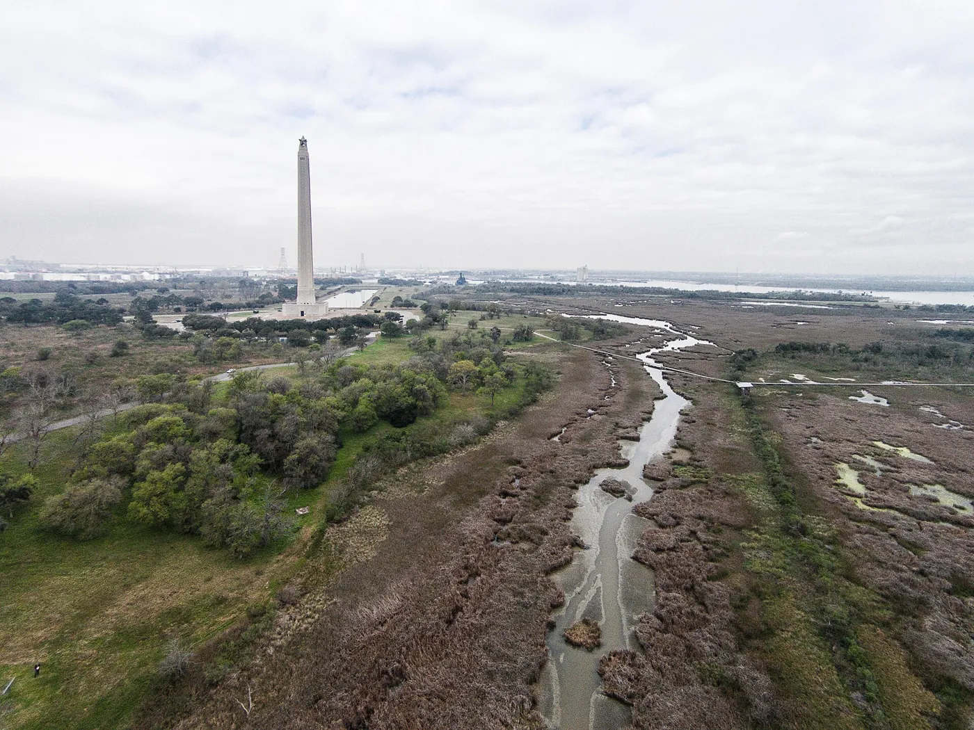 An aerial view of marsh and a winding creek on flat coastal prairie, with the San Jacinto Monument and the Houston Ship Channel in the distance
